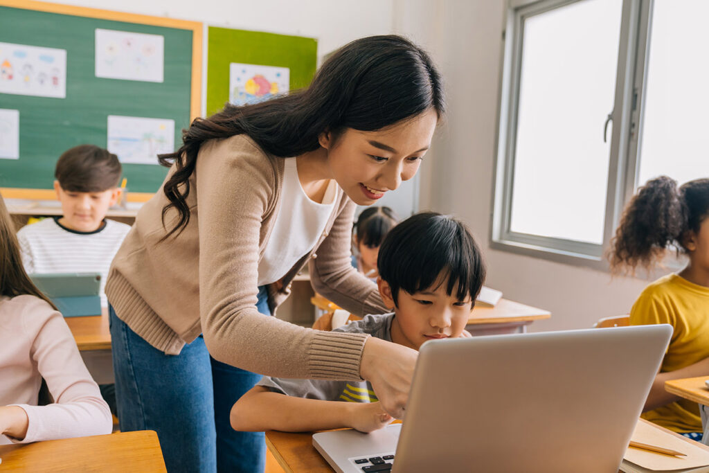 Asian teacher helping elementary student boy with laptop in computer classroom. Information Technology class in primary school concept.